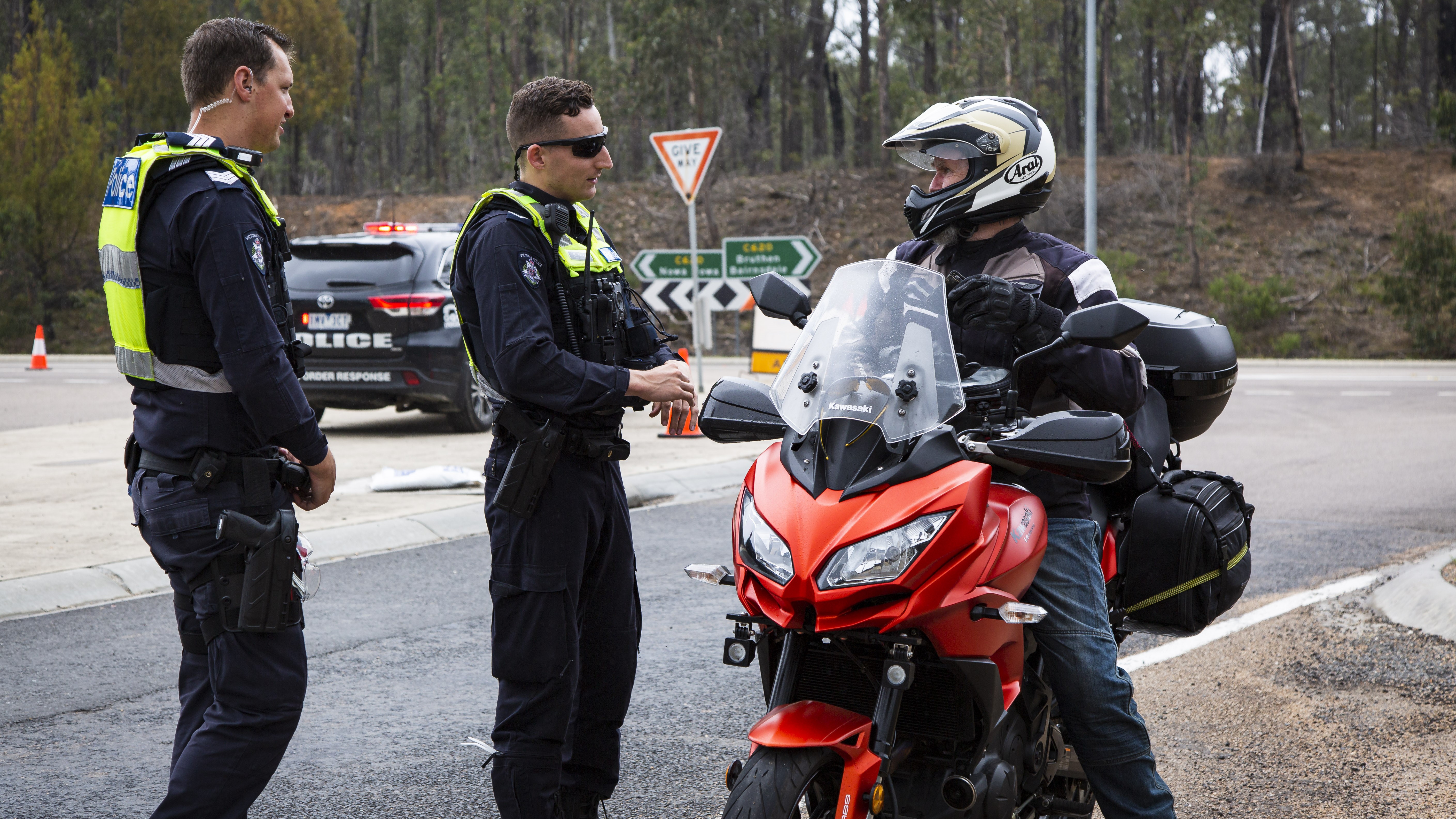 Two officers are stopped on the side of the road, talking to a person sitting on their motorcycle, wearing a helmet and bike gear.