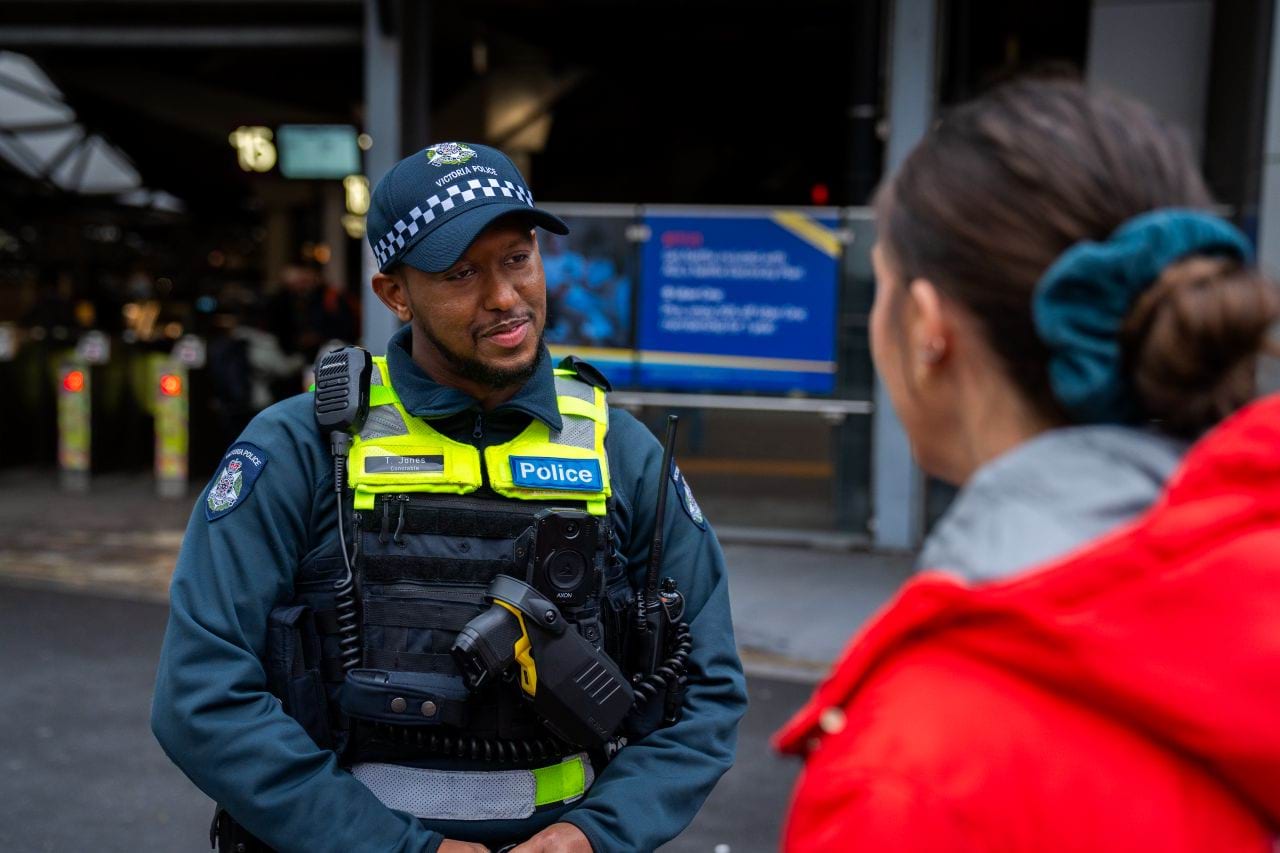Police officer talking to a woman on the street who is wearing a bright red jacket. He is attentively listening to what she's saying, with his head on the side. 