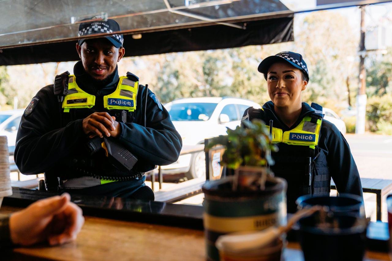 Two officers are standing outside an open cafe window. looking in at a working. They are both smiling and we see them from the cafe workers point of view. 