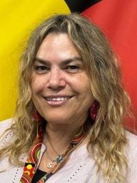 Selina Grizos with long wavy hair, wearing a light-coloured top, earrings and a lanyard with a patterned strap. A yellow, red and black background is visible behind her.